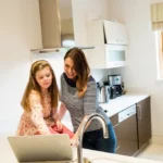 A woman and a young girl smiling while looking at a laptop together in a bright, modern kitchen.