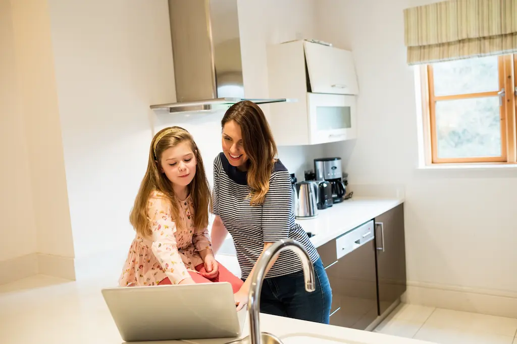 A woman and a young girl smiling while looking at a laptop together in a bright, modern kitchen.