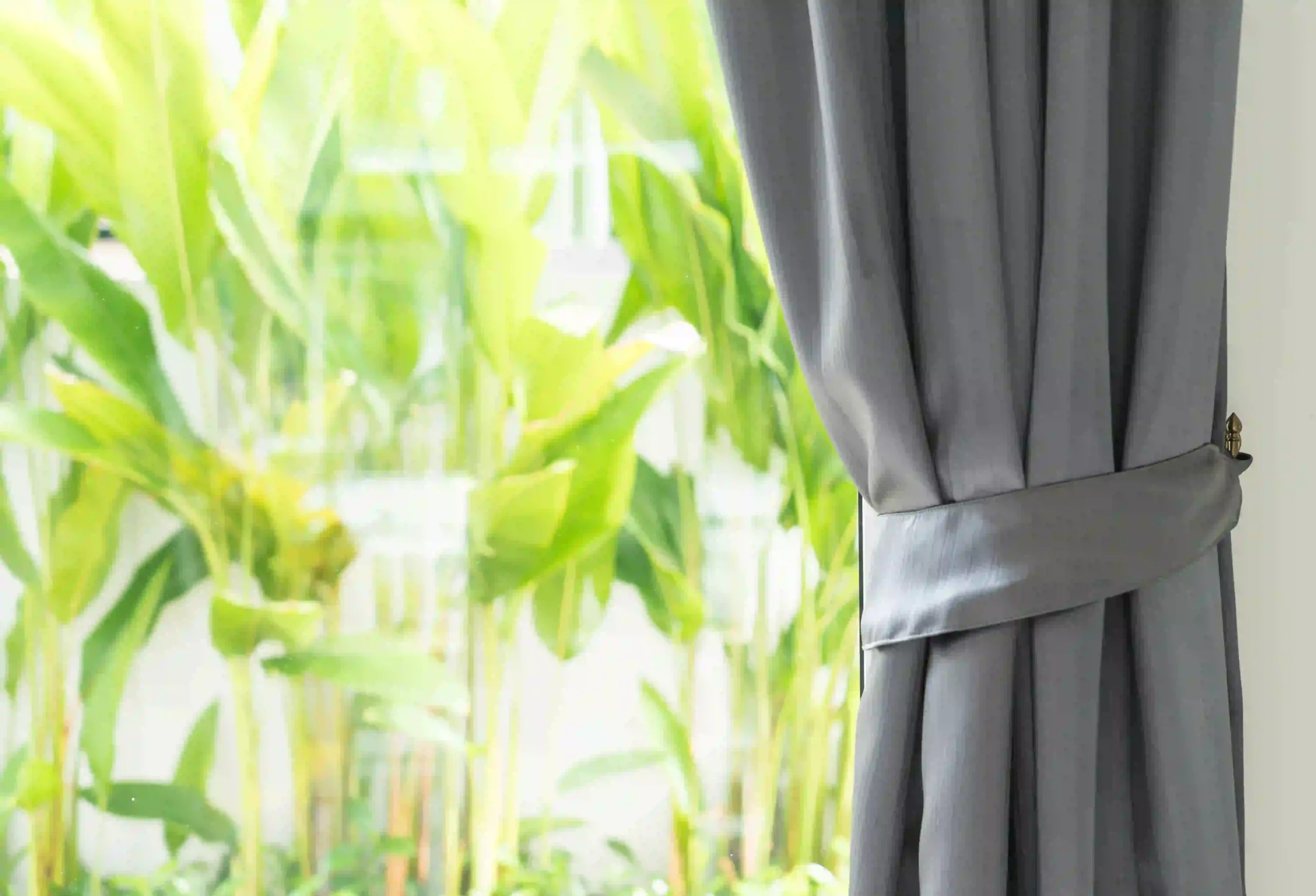 The image shows a close-up view of a grey curtain neatly tied back with a matching fabric band. The curtain is hanging beside a window, allowing bright natural sunlight to enter the room.