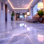 Low-angle view of a glossy marble tile floor in a modern hallway with warm ceiling lights, indoor plants, and elegant seating in the background.
