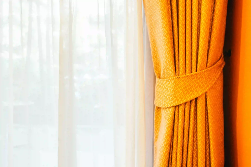 This curtain image shows a beautifully styled window with two layers: a soft white sheer curtain in the background and a rich golden drape tied neatly to the side. The warm color and elegant fabric create a luxurious and welcoming atmosphere.