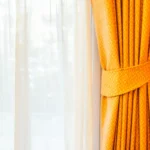 This curtain image shows a beautifully styled window with two layers: a soft white sheer curtain in the background and a rich golden drape tied neatly to the side. The warm color and elegant fabric create a luxurious and welcoming atmosphere.