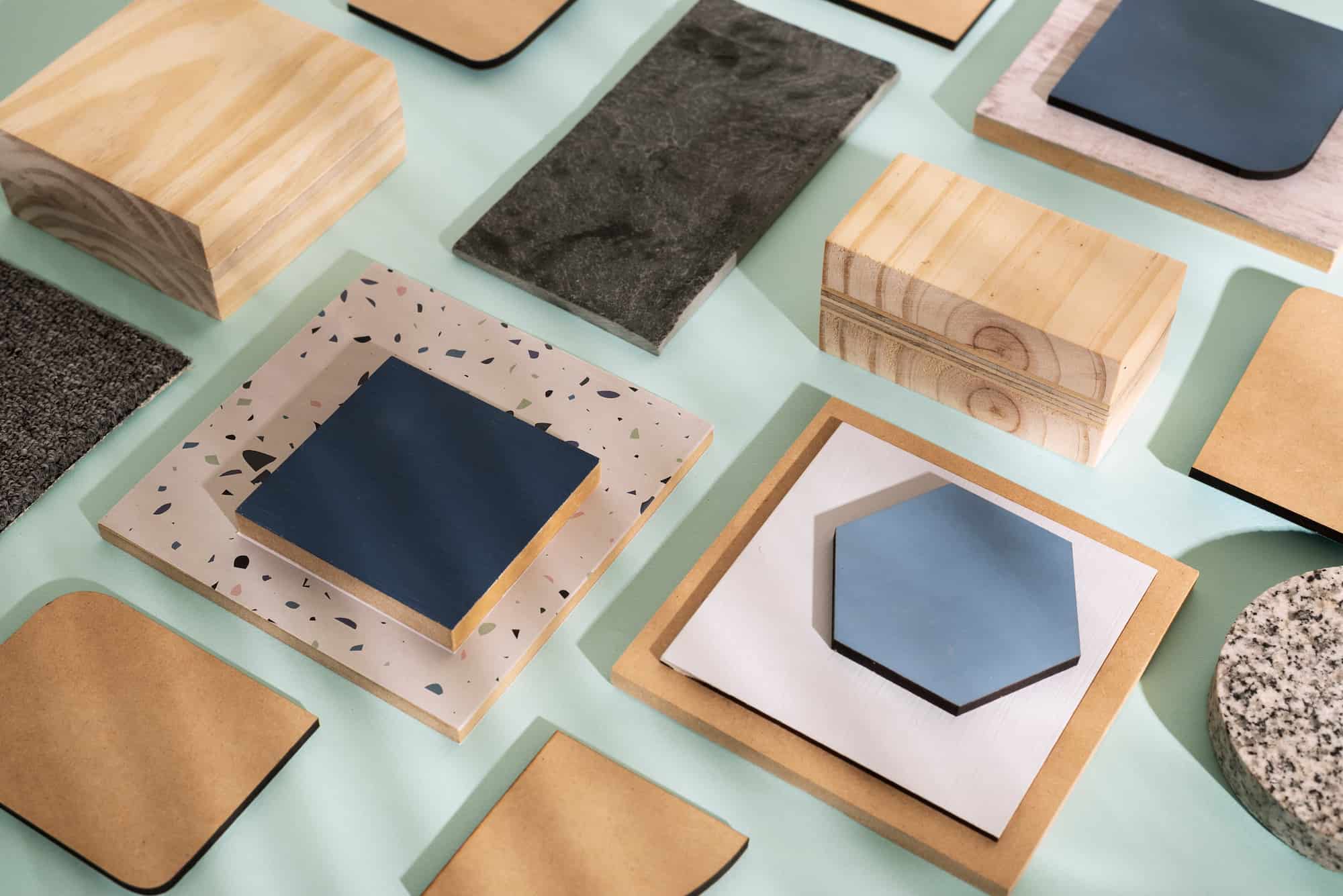 Top view of assorted material samples including wood blocks, MDF boards, terrazzo tile, carpet swatch, stone slab, and blue ceramic tiles arranged on a pastel green background.