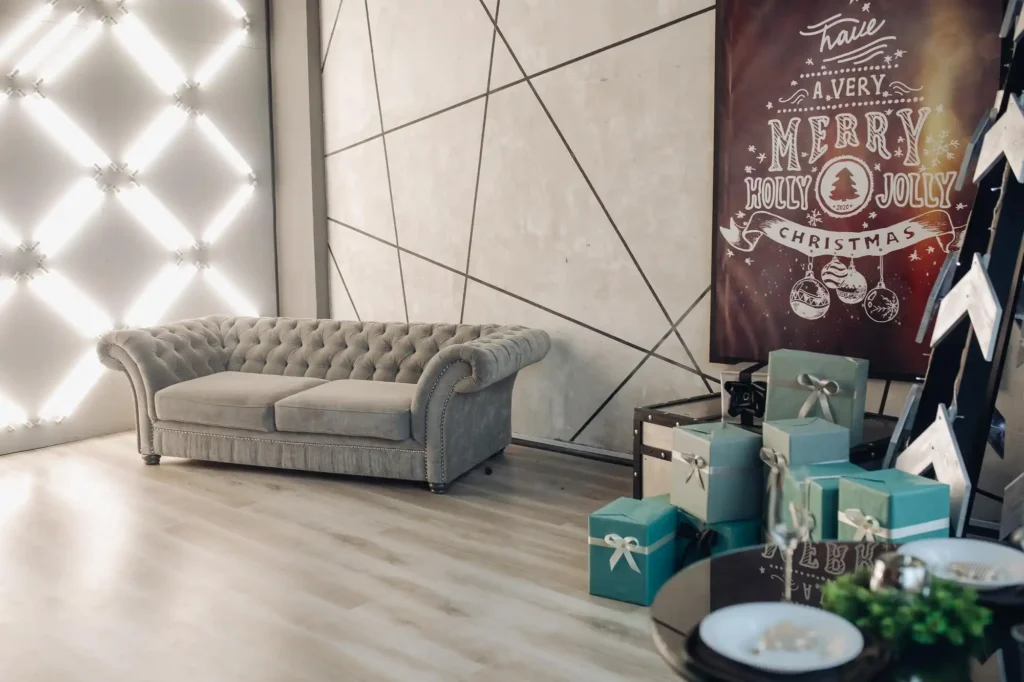 Modern living room with a grey tufted sofa, geometric wall design, LED light accents, and Christmas decorations including gift boxes and a festive “Merry Holly Jolly Christmas” poster.