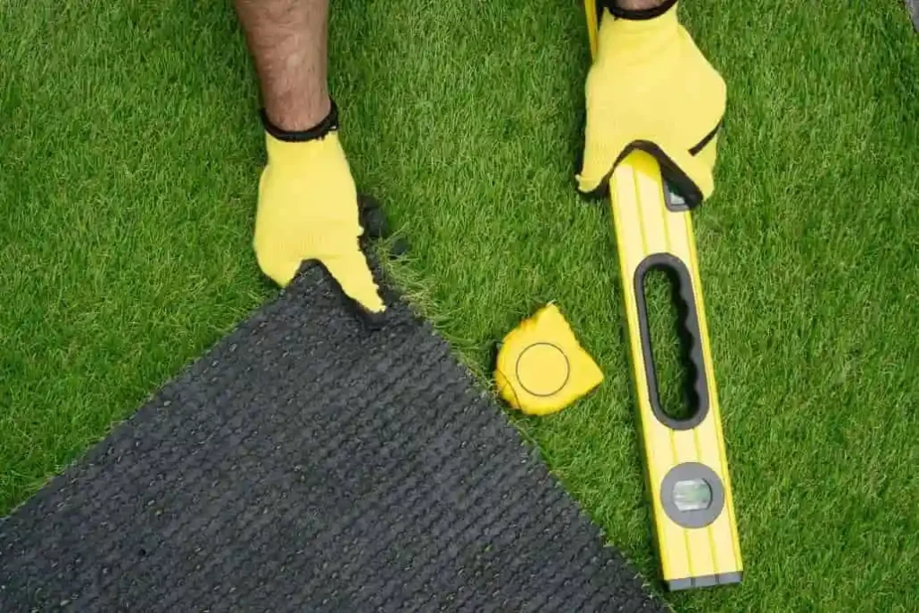 Person installing artificial grass using a level and measuring tape while aligning turf edges.