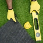 Person installing artificial grass using a level and measuring tape while aligning turf edges.