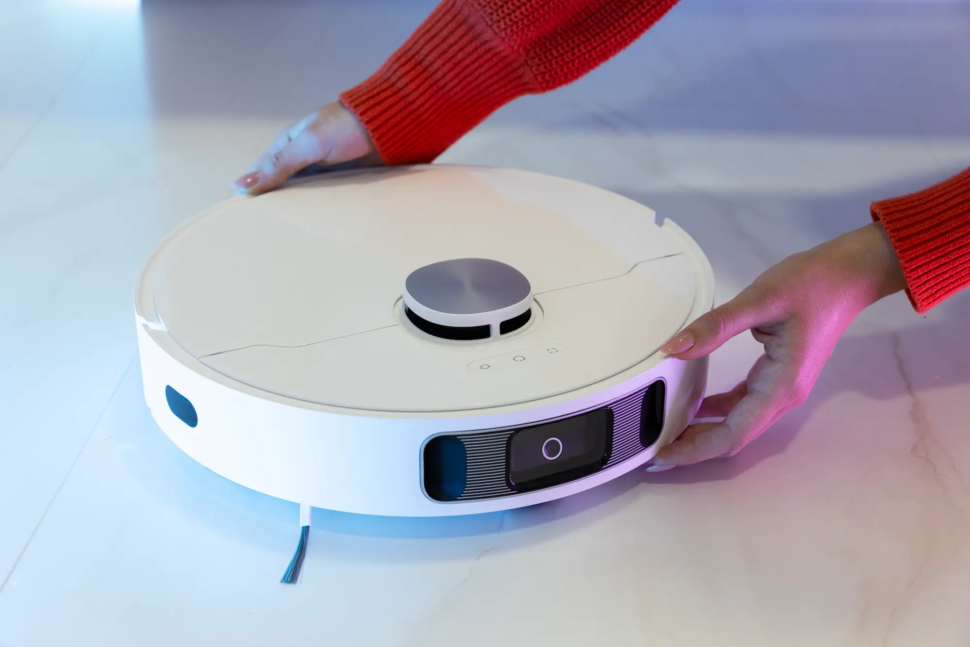 Alt text: A person in a red sweater adjusts a white robotic vacuum cleaner with a top-mounted sensor and front-facing camera on a light-colored floor.