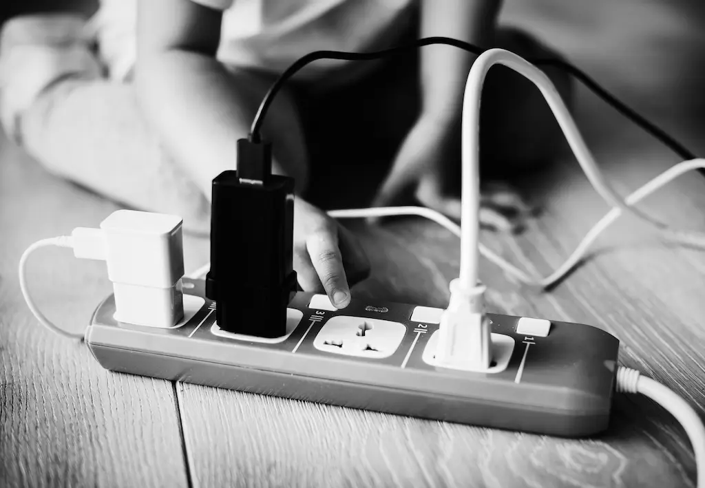 Alt text: Black-and-white photo of a child’s hand reaching toward a power strip with multiple chargers plugged in, placed on a wooden floor.