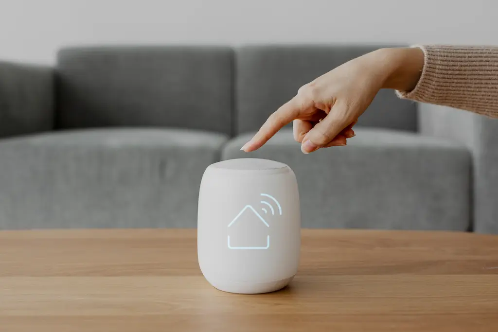 Hand pressing a smart home control device with a house and Wi-Fi icon on a table in a modern living room.