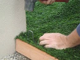 A person securing the edge of artificial grass with a hammer and nail along a wooden border during turf installation.