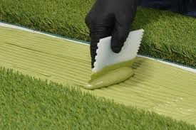 A gloved hand spreading adhesive with a notched trowel to join two sections of artificial grass during turf installation.