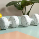 A clean and inspiring home gym setup featuring essential DIY fitness accessories arranged neatly in a cozy corner of a room. The space includes a pair of dumbbells resting on a rubber mat, a rolled-up yoga mat leaning against the wall, and colorful resistance bands hanging from a simple wall hook