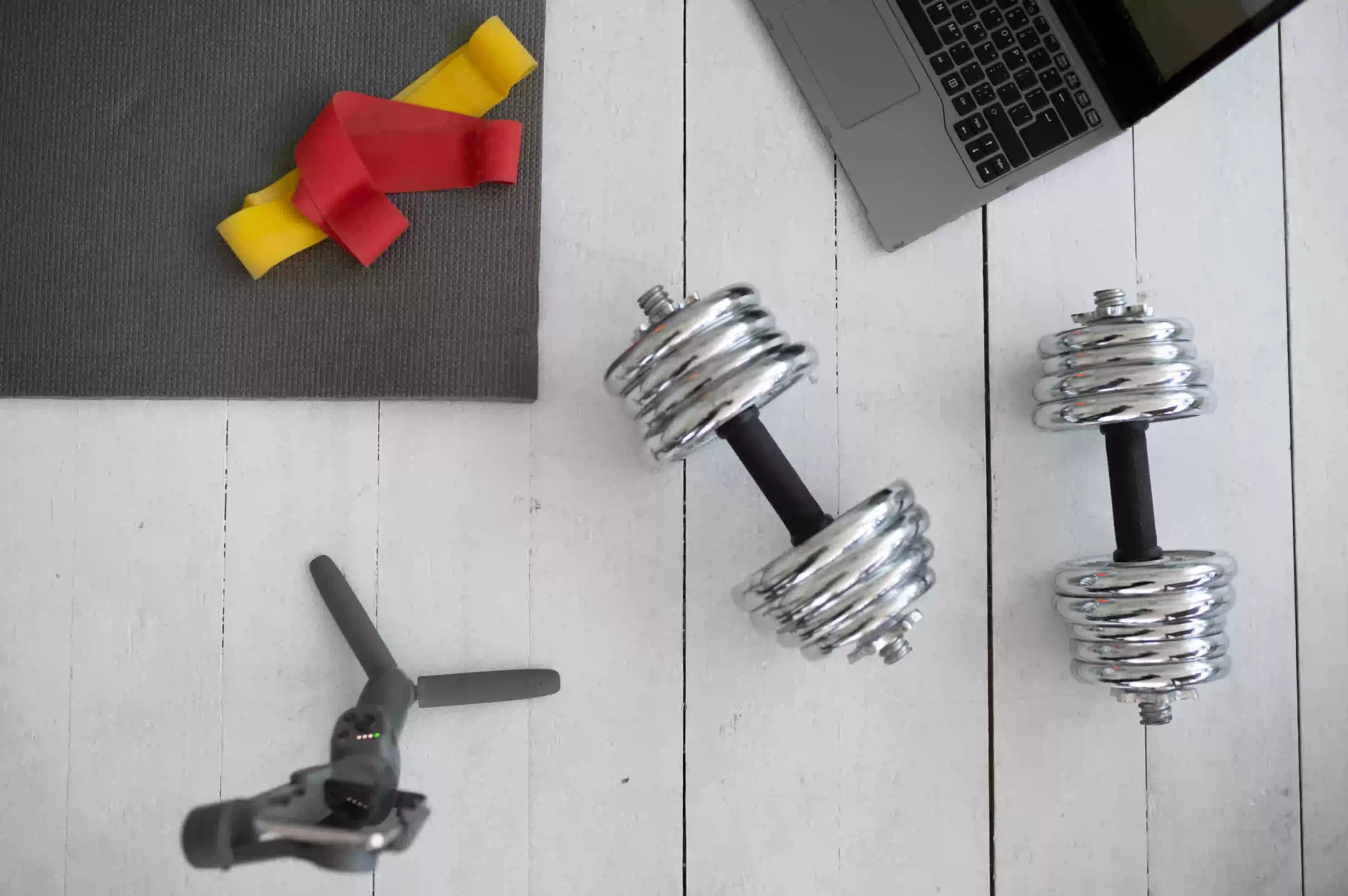 A clean and inspiring home gym setup featuring essential DIY fitness accessories arranged neatly in a cozy corner of a room. The space includes a pair of dumbbells resting on a rubber mat, a rolled-up yoga mat leaning against the wall, and colorful resistance bands hanging from a simple wall hook