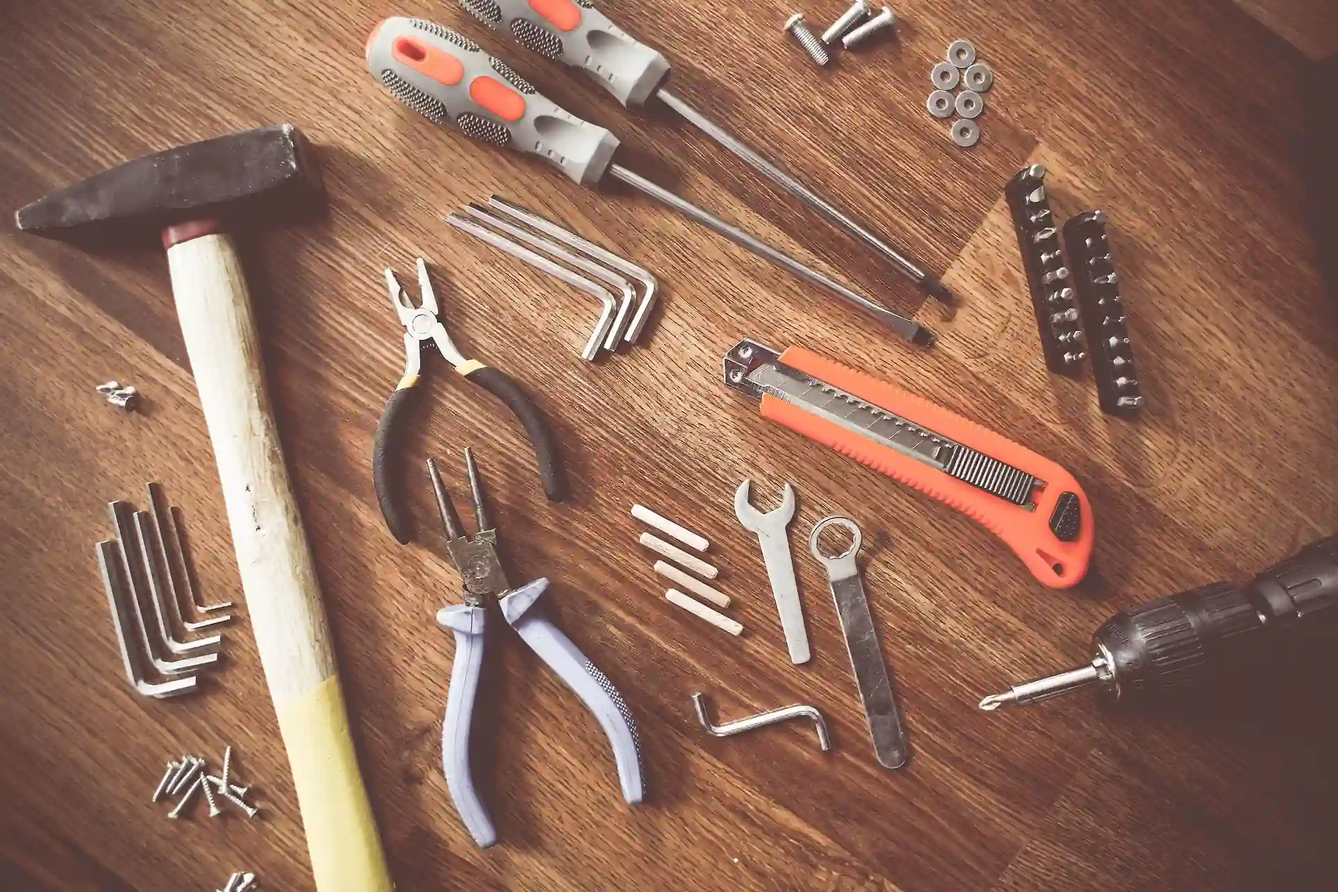 Assorted DIY hand tools laid out on a wooden surface, including a hammer, pliers, screwdrivers, hex keys, wrenches, a utility knife, a drill bit, and various screws and bolts.