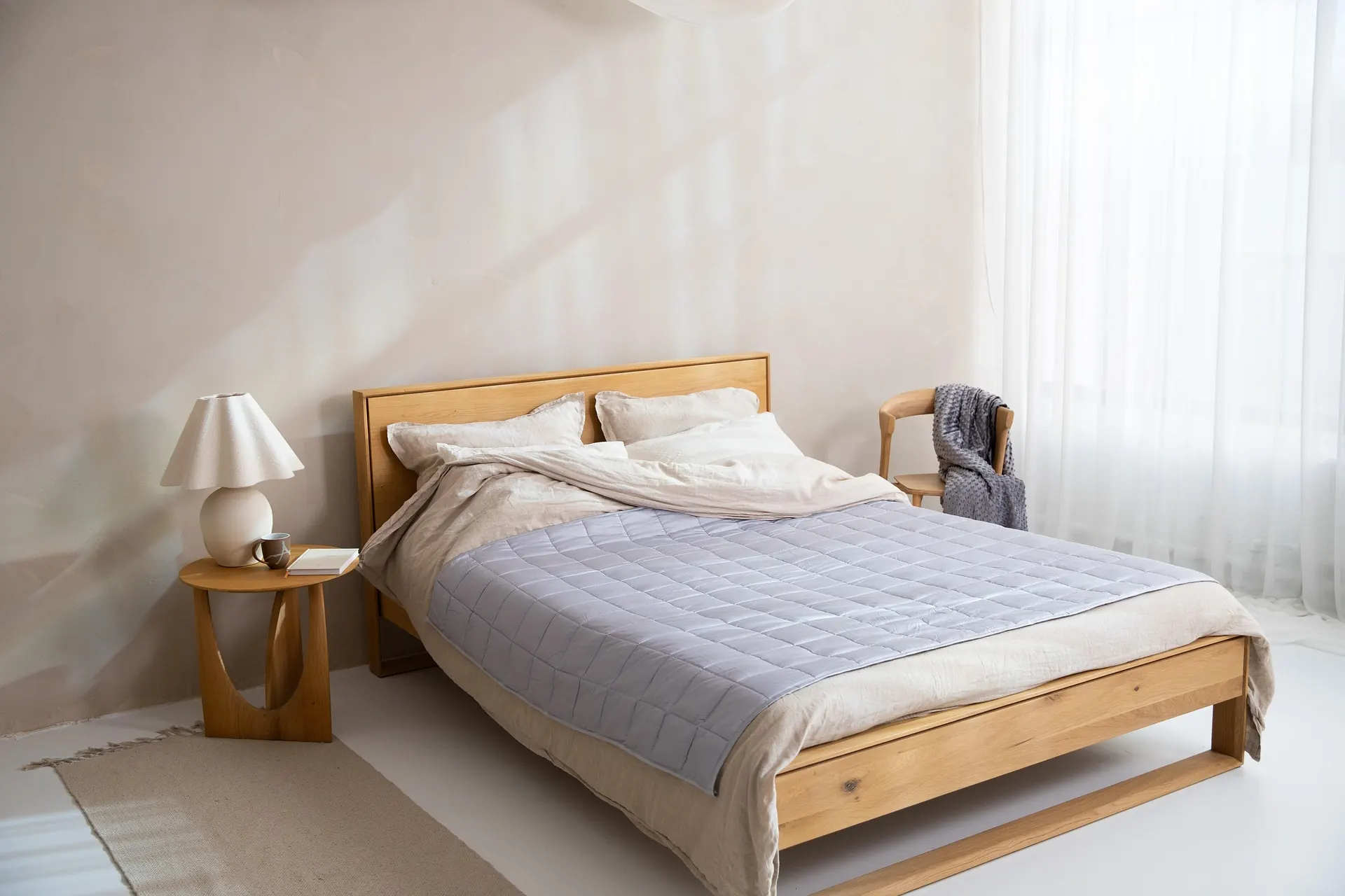 A minimalist bedroom featuring a solid wood bed frame with a simple headboard, dressed with beige linen bedding, cream pillows, and a light grey quilted blanket, next to a round wooden nightstand with a lamp and a mug.